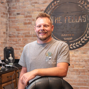 Man standing behind a barber chair with The Fellas logo on the wall behind.